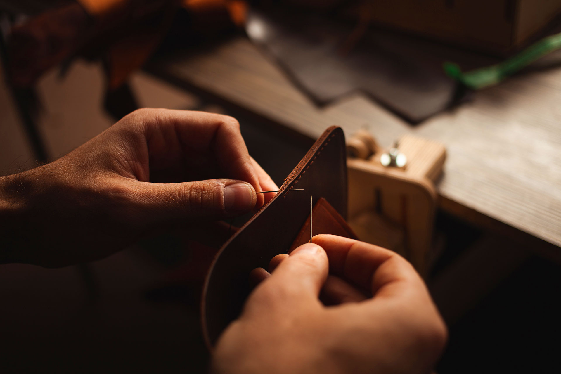 Hands hand-stitching a leather wallet with needle and thread on a workbench
