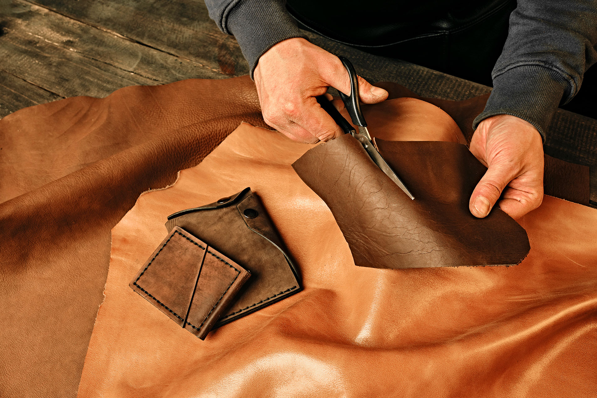 Leather craftsman cutting brown hide with scissors next to a handmade wallet